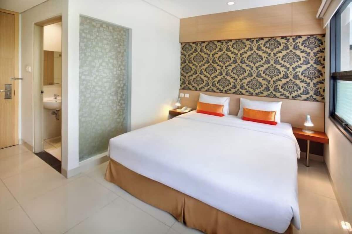 A hotel room with a large bed, a nightstand, a bedside table, a bathroom with a toilet and sink, and a patterned wall.