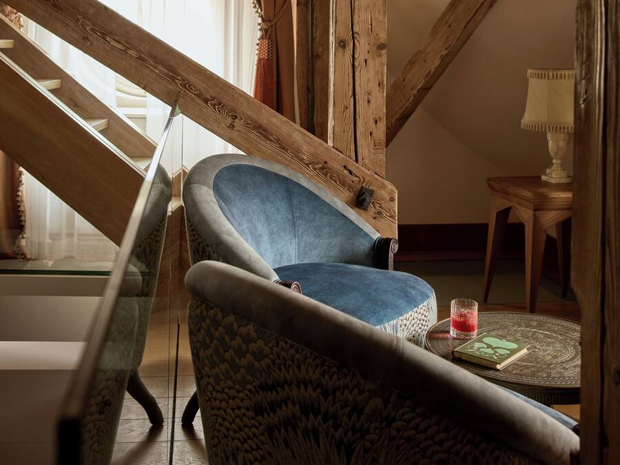 Una habitación acogedora con una escalera de madera, un sillón azul, un sofá estampado y una mesita con un libro y un vaso.