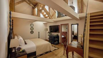 A bedroom with a bed, bedside table, lamp, and a TV. There is a wooden desk with a chair and a mirror. A staircase leads to an upper level.