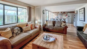 A spacious living area with leather sofas, a wooden coffee table, and large windows offering a view of the garden.