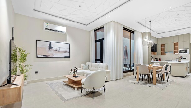 A modern living room with a sofa, coffee table, and dining area with chairs. There is a kitchen with a microwave and cabinets.