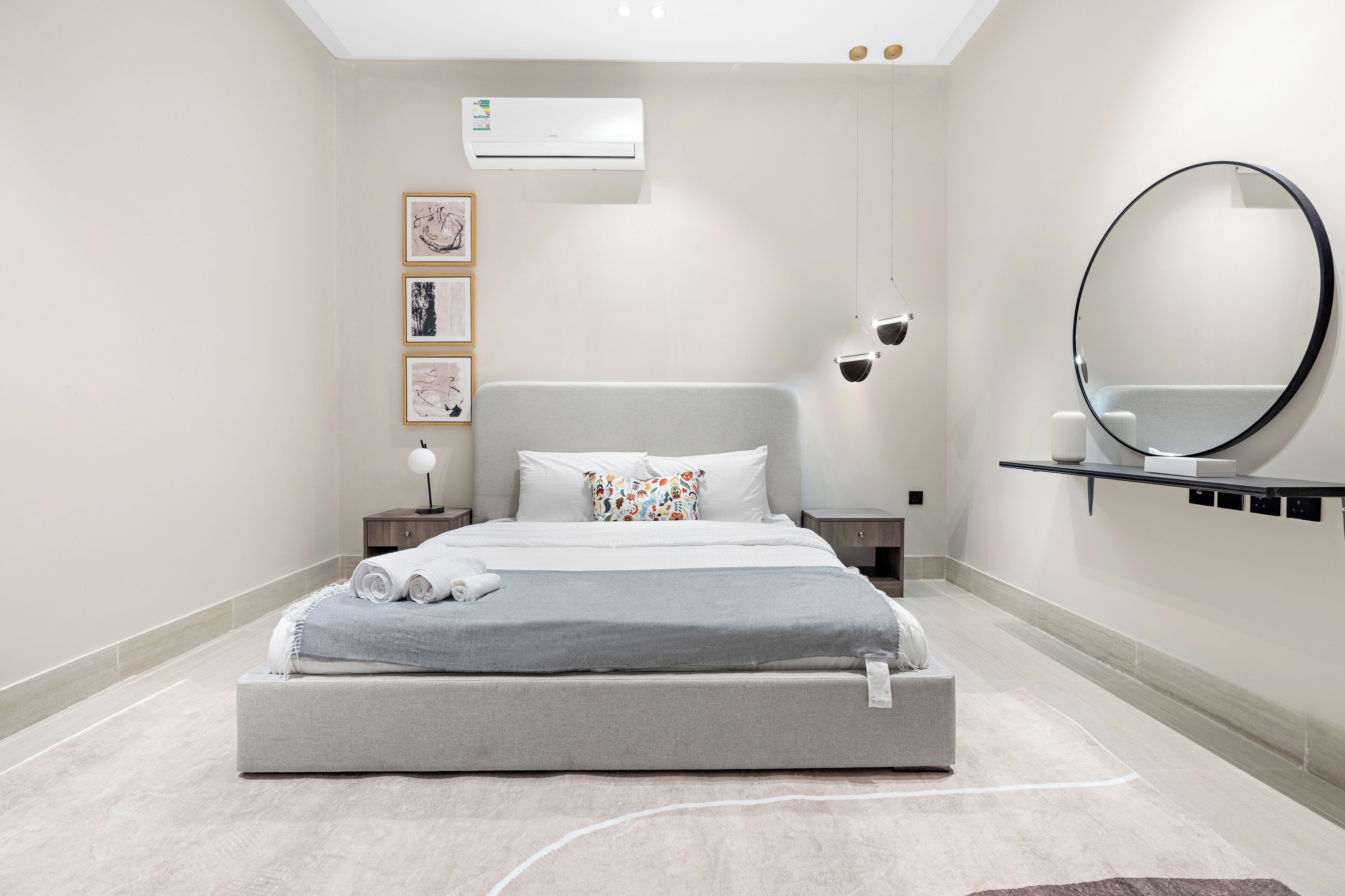 Une chambre moderne avec un grand lit, une table de chevet avec une lampe, un climatiseur fixé au mur et un miroir rond placé au-dessus d’une étagère.