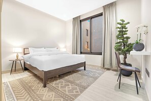 A modern hotel room with a large bed, a small side table with a lamp, a potted plant, and a view of the outside through a window. - Silkhaus Ramz 49 (Riyadh)
