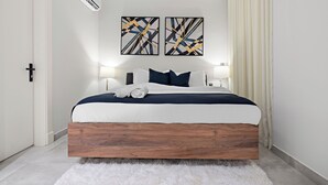 A modern bedroom with a wooden bed frame, white bedding, and two abstract paintings on the wall. - Silkhaus Sayl 25 (Riyadh)