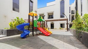 A playground with slides and climbing structures, surrounded by a courtyard with tiled flooring and greenery. - Silkhaus Sayl 25 (Riyadh)
