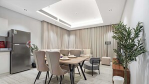 Et moderne køkken med et indbygget køleskab, et spiseområde med et træbord og stole, samt en opholdsstue med sofa og gardiner.