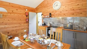 Interior de madeira com uma mesa de jantar para quatro pessoas, área de cozinha com fogão e pia, e um relógio de parede.