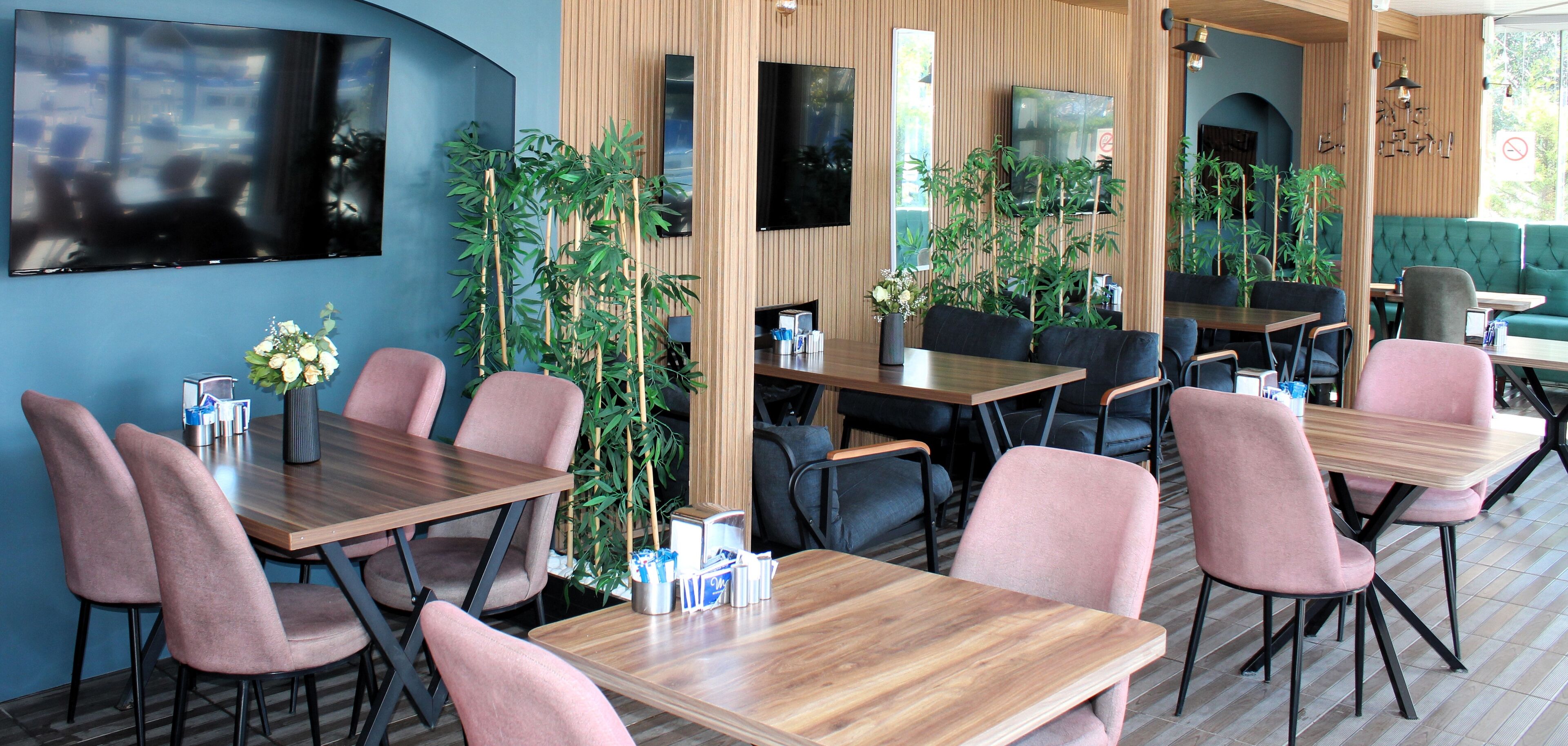 A modern restaurant with wooden tables and chairs, large windows, and indoor plants.