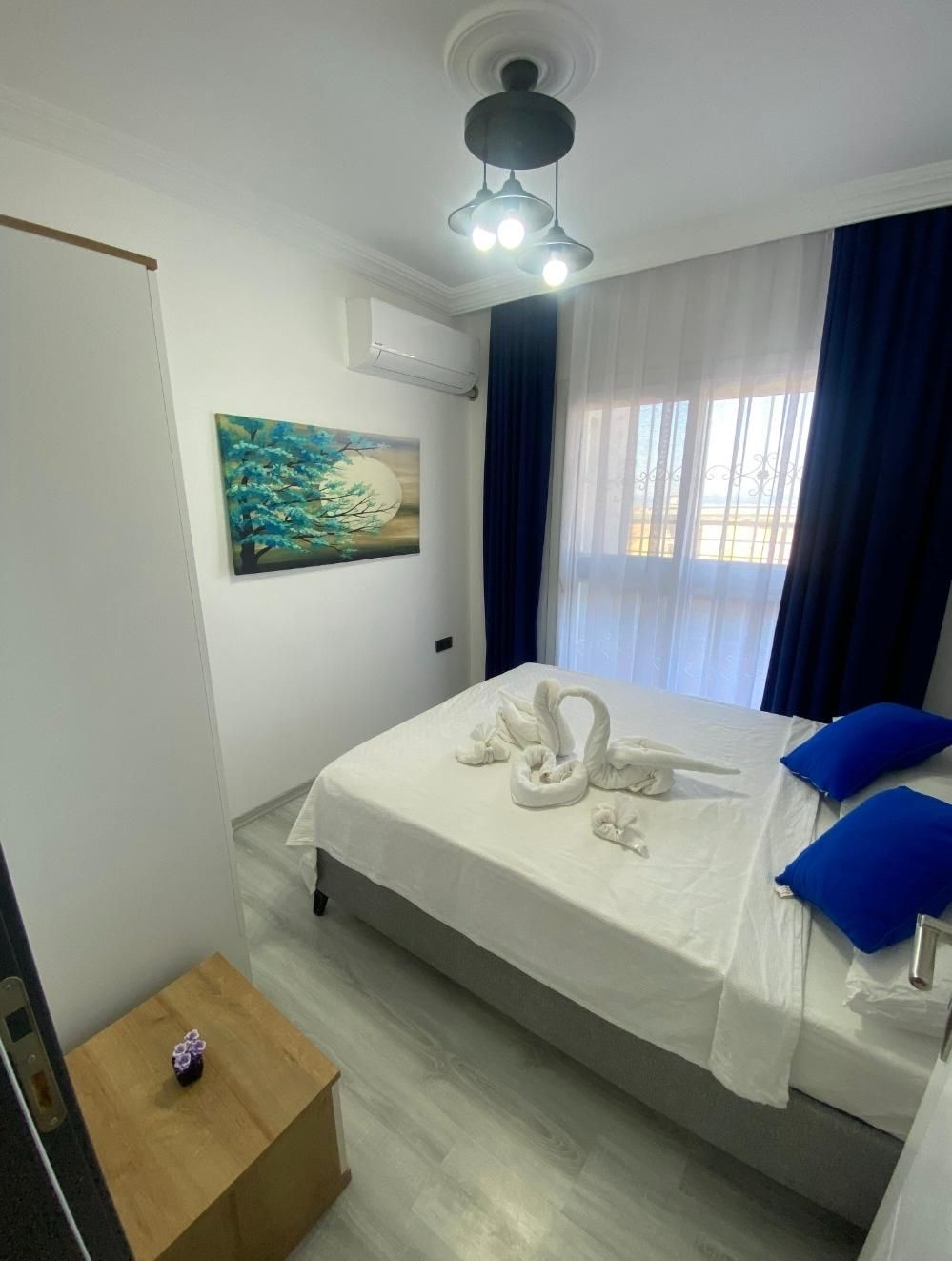 A hotel room with a bed, a painting, a ceiling light, and a small wooden table.