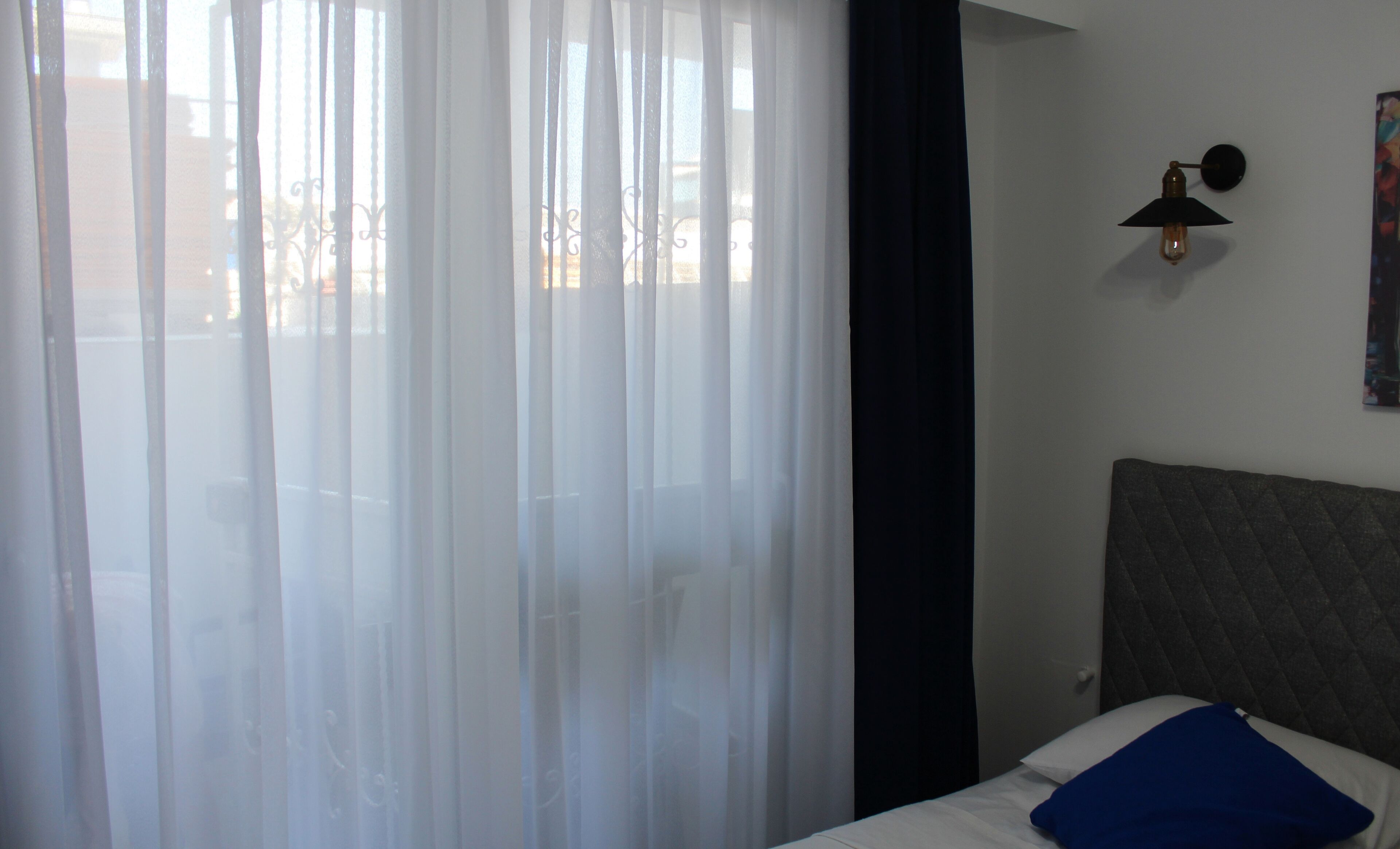 Quarto de hotel com cama, mesa de cabeceira, luminária na parede e cortinas translúcidas.