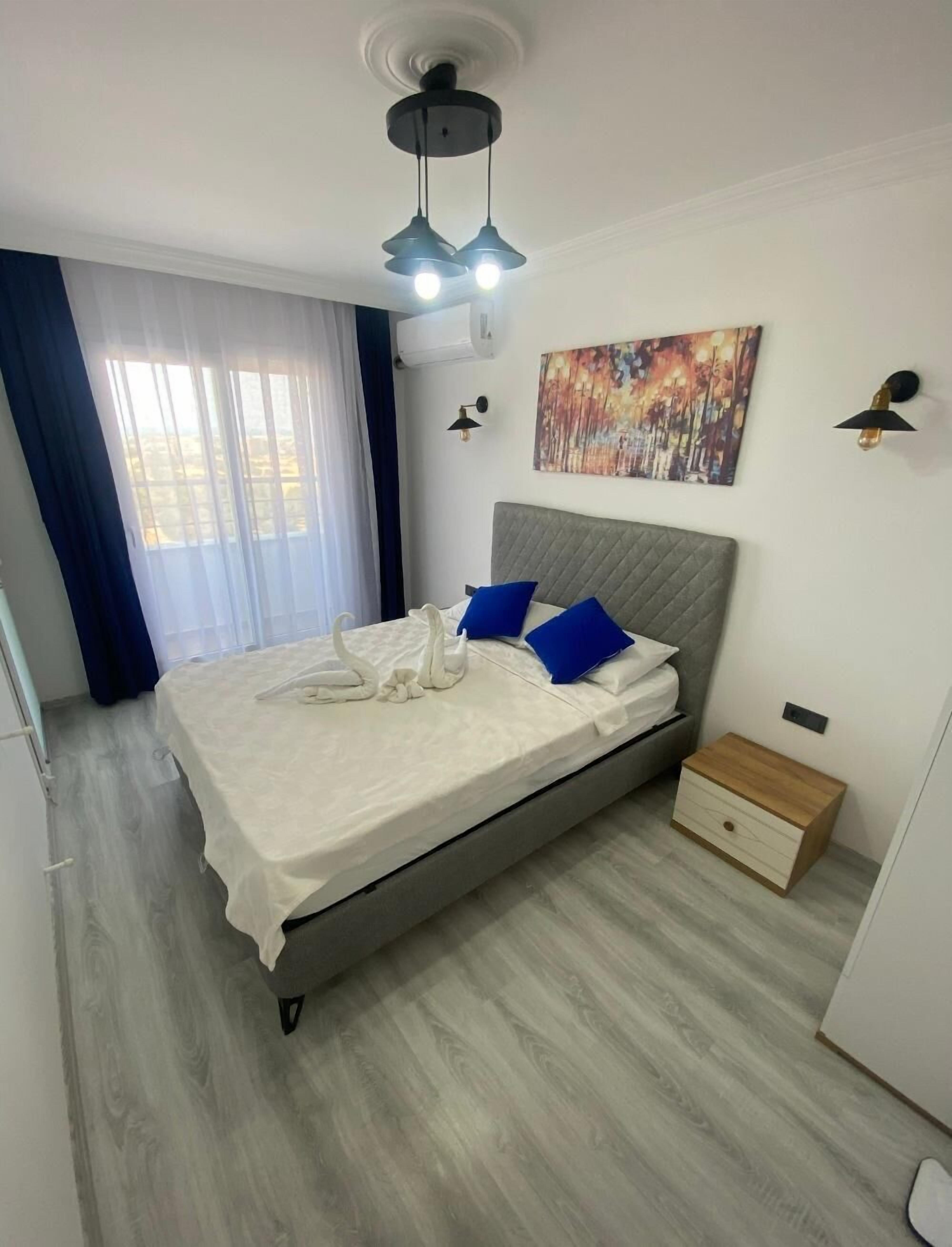 Un dormitorio con cama, mesita de noche, lámpara de pared, un cuadro y ventana con cortinas.
