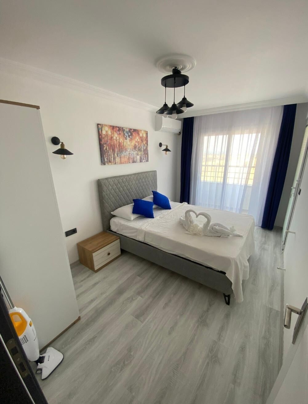 Una habitación de hotel moderna con una cama, una mesita de noche, un ventilador de techo, una lámpara de pared, un cuadro y una ventana con cortinas.