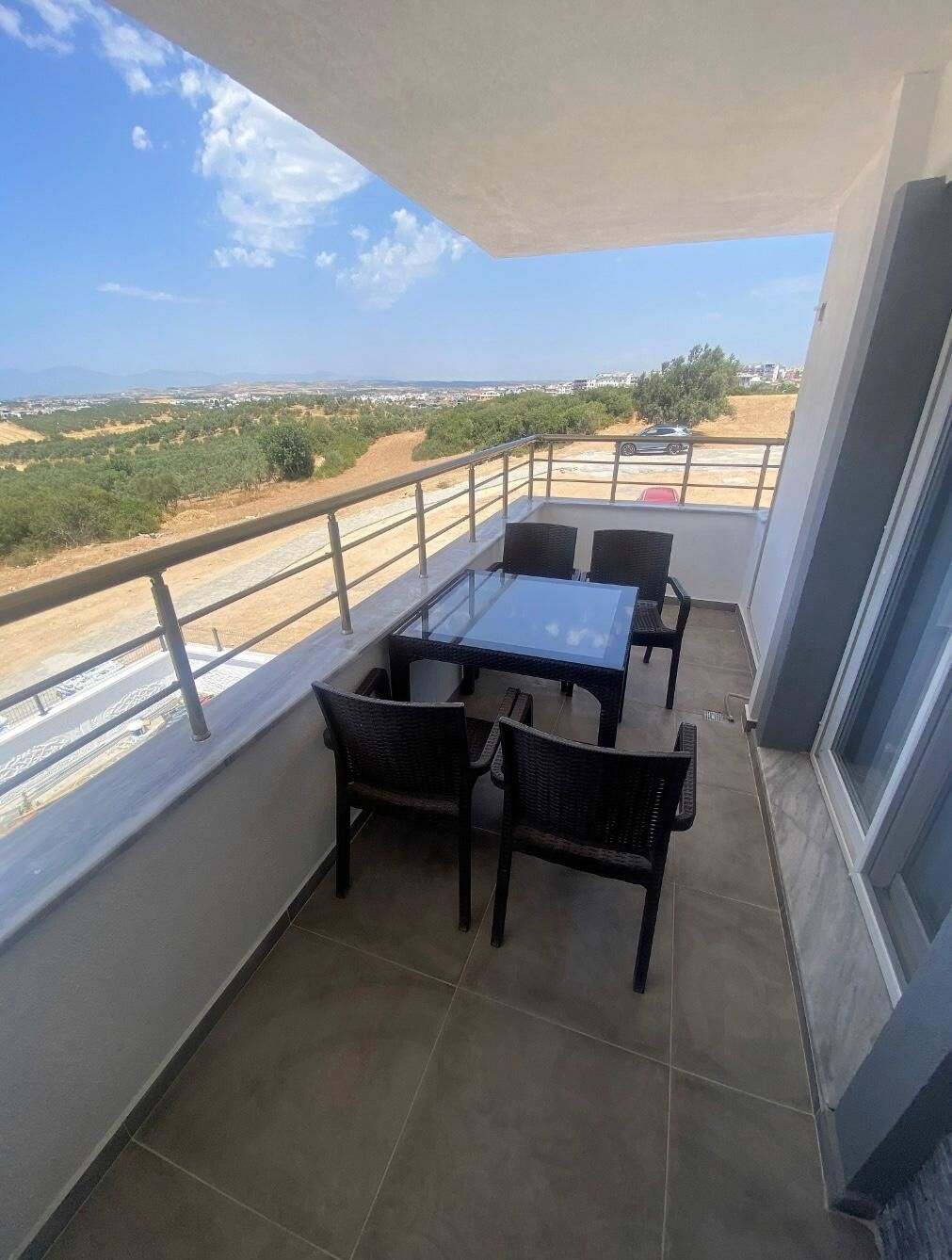 Un balcon doté d’une table et de chaises en verre, offrant une vue sur une route et sur la verdure.