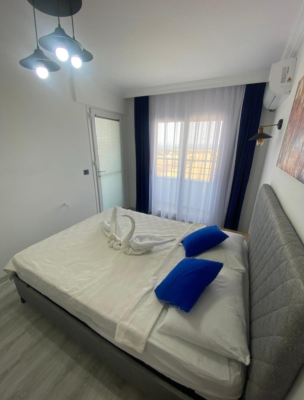 A neatly made bed with white and blue pillows, a white duvet, and a towel shaped like a swan. There is a ceiling light fixture above the bed.