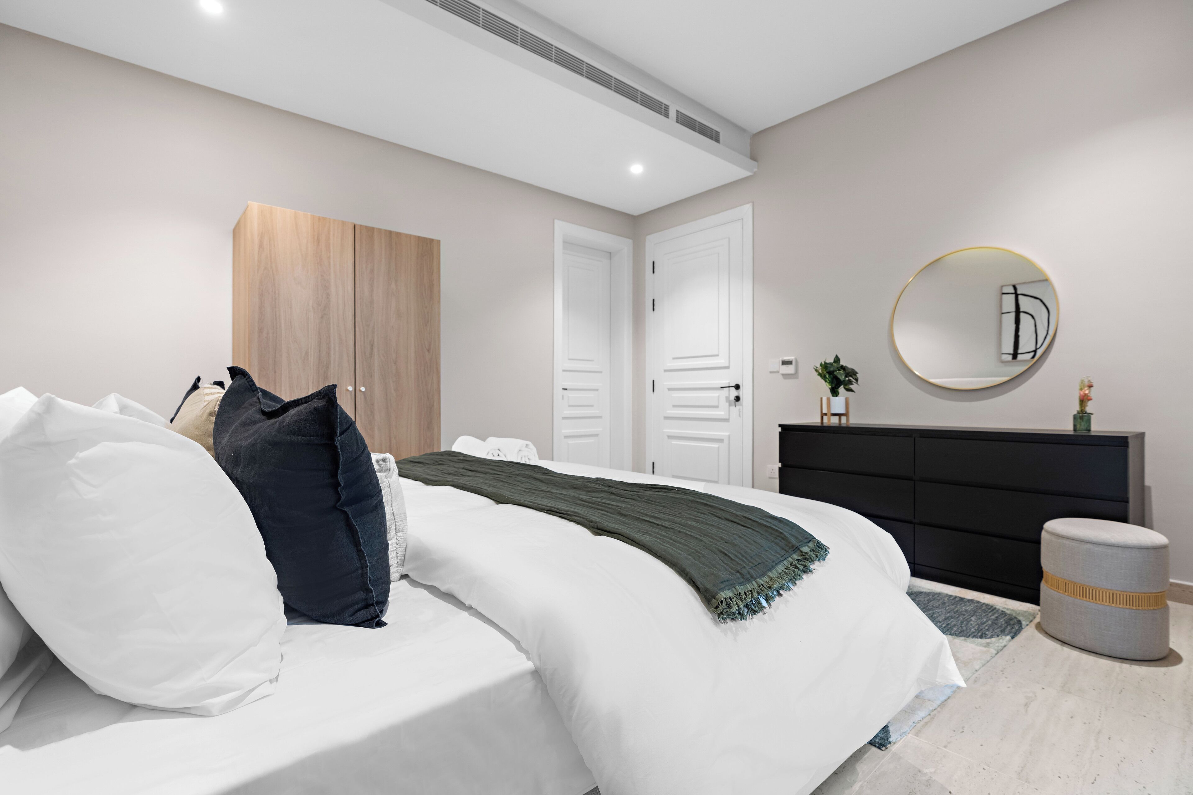 A modern bedroom with a bed, white bedding, a dark headboard, a wooden wardrobe, a black dresser, a round mirror, and a small round ottoman.