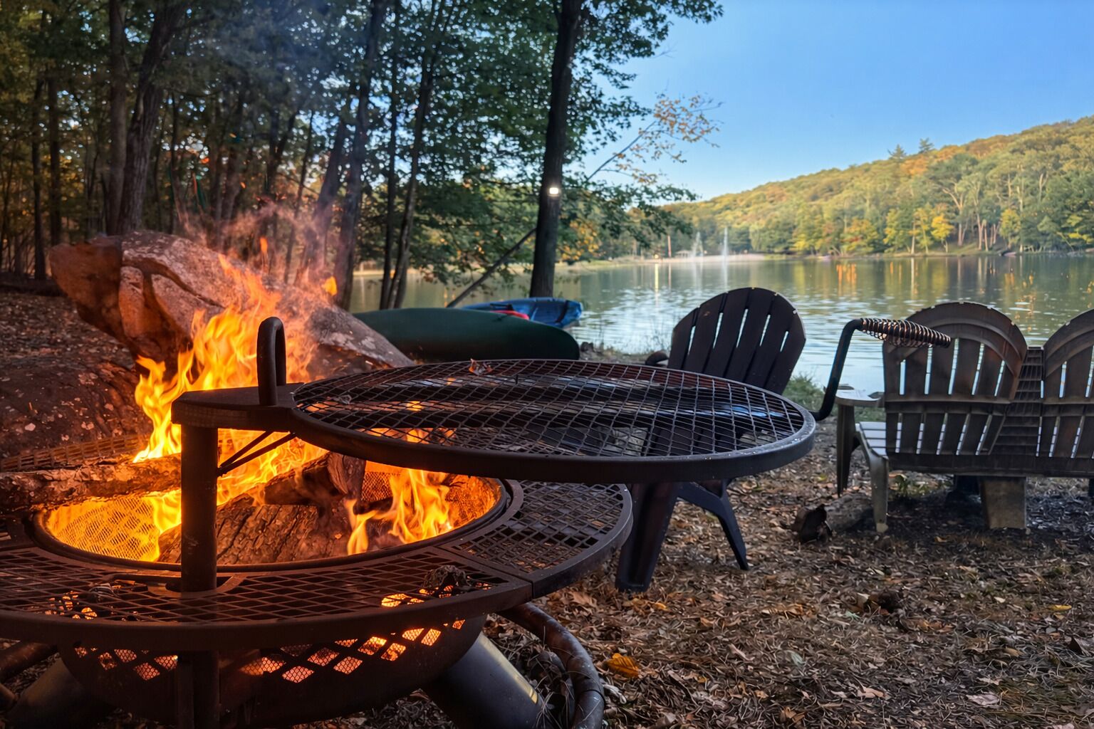 A campfire with a metal grill, two metal chairs, and a bench near a lake surrounded by trees.