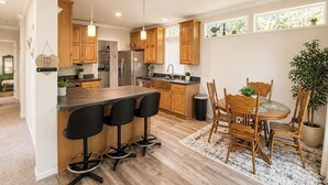 Una cocina moderna con armarios de madera, una isla central con taburetes de bar, una zona de comedor con mesa y sillas de madera, y vistas al exterior a través de amplias ventanas.