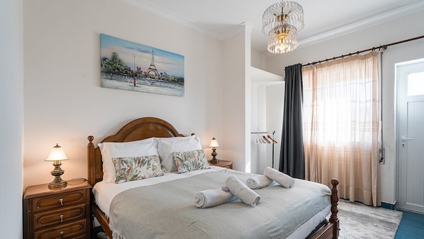 Een net opgemaakt bed met een hoofdbord, twee nachtkastjes met lampen, een schilderij van de Eiffeltoren en een groot raam met gordijnen.