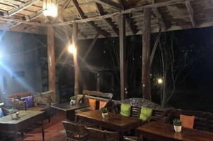 Un área de comedor al aire libre con mesas y sillas de madera, techo de paja y vista al cielo nocturno.