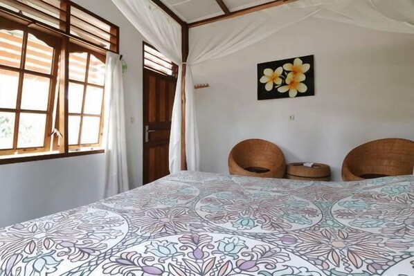 A room with a bed, two wicker chairs, a window with curtains, and a wall with a flower painting.