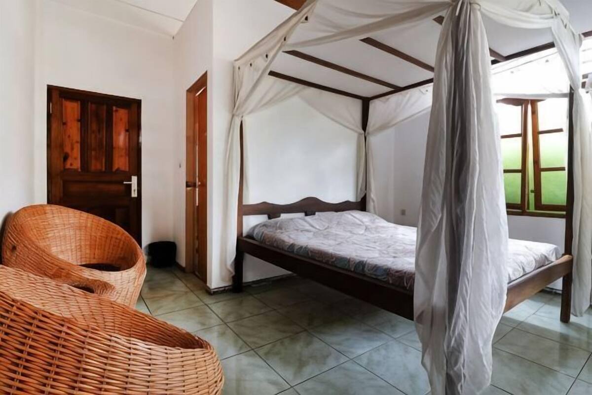 A room with a bed under a canopy, a wicker chair, and a door.