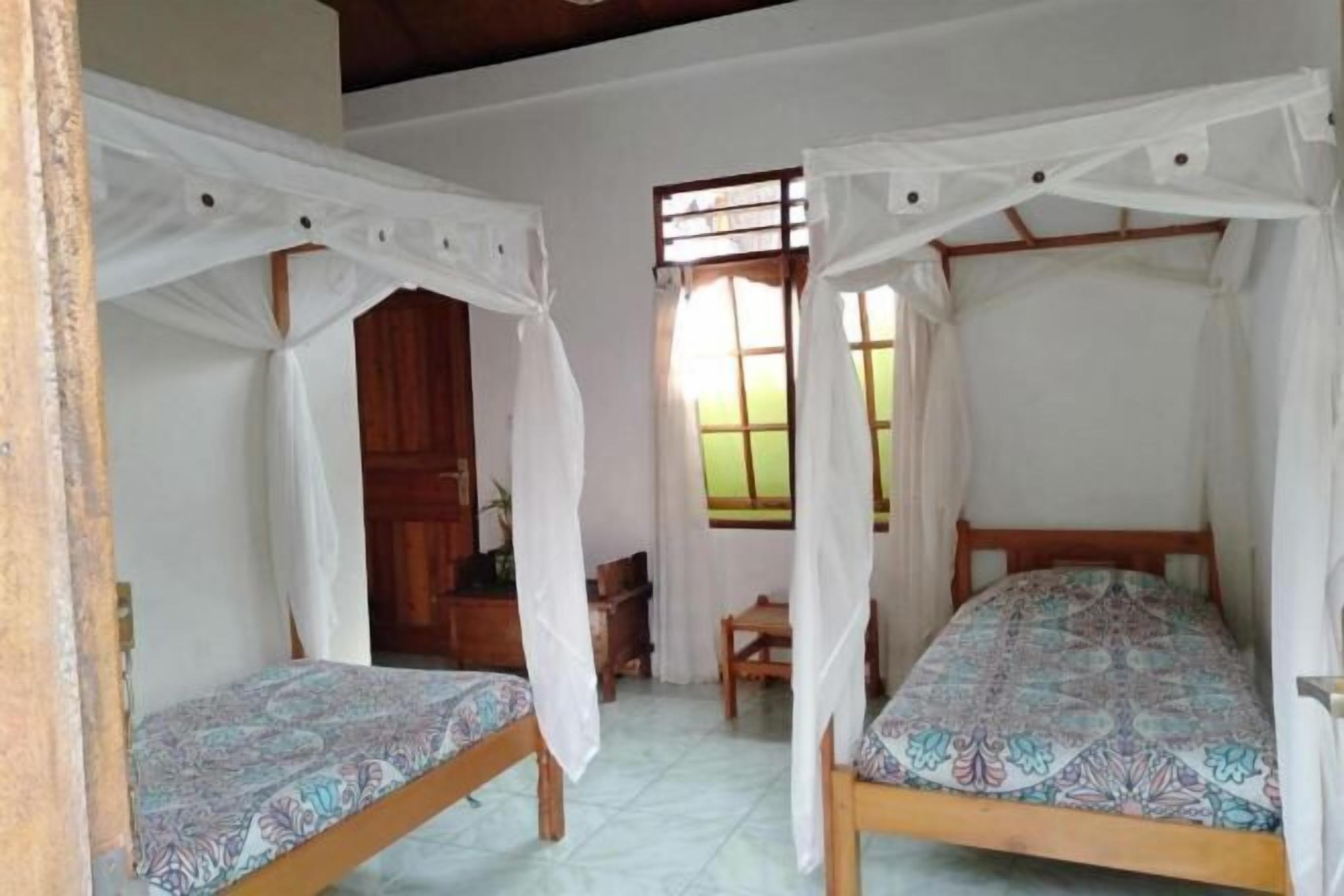 Quarto com duas camas de solteiro, cada uma com dossel, porta de madeira, uma mesinha e uma janela com cortinas.