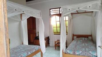 Quarto com duas camas de solteiro, cada uma com dossel, porta de madeira, uma mesinha e uma janela com cortinas.