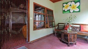 A room with a wooden ceiling, a large window with curtains, a wooden bench, a round wooden table with a vase, and a wall with a flower painting. - Bagus Bay Homestay (Tuktuk Siadong)