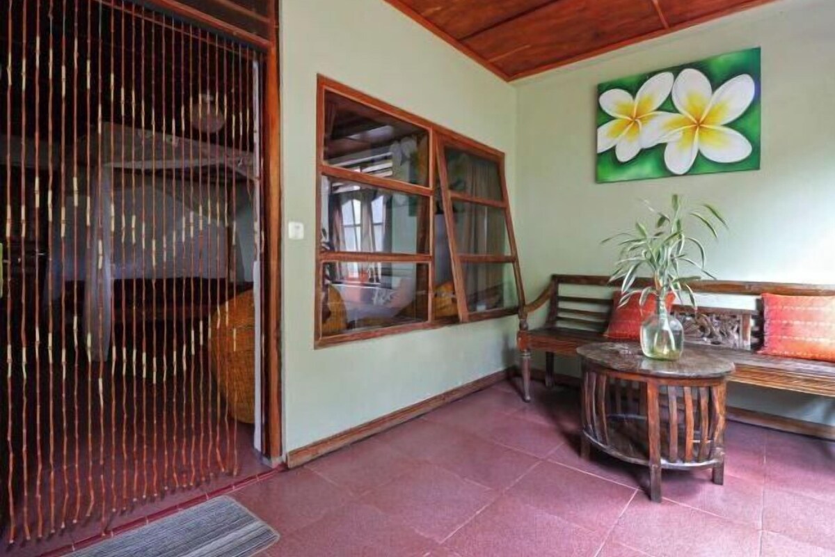 A room with a wooden ceiling, a large window with curtains, a wooden bench, a round wooden table with a vase, and a wall with a flower painting.