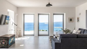 Un salon moderne avec un canapé gris, une commode en bois et un trépied. Trois grandes fenêtres offrent une vue sur la mer.