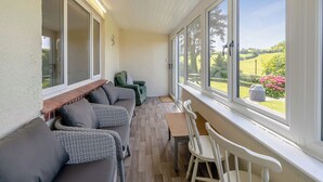 Une terrasse couverte, aménagée avec un canapé, des fauteuils et une table, d’où l’on peut admirer un paysage verdoyant, parsemé d’arbres et de fleurs.