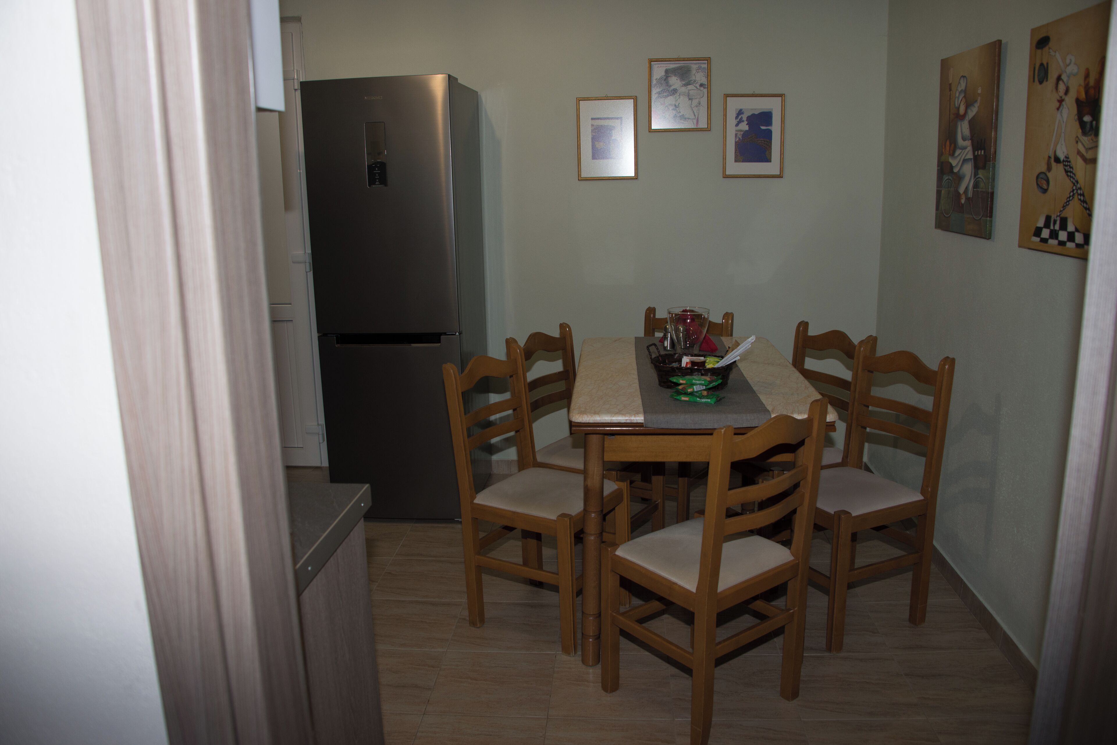 A compact kitchen with a refrigerator, wooden dining table, and four chairs. There are framed pictures on the wall.