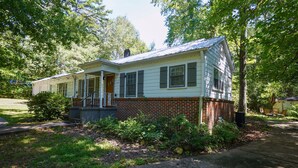 A two-story house with a brick foundation and white siding, surrounded by lush greenery and trees. - Pine Haven - Large Group - Baby & Pet friendly (Birmingham)