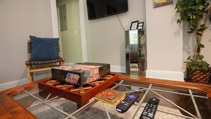 A living room with a glass coffee table, a sofa with a blue cushion, a television mounted on the wall, and a potted plant. - Pine Haven - Large Group - Baby & Pet friendly (Birmingham)