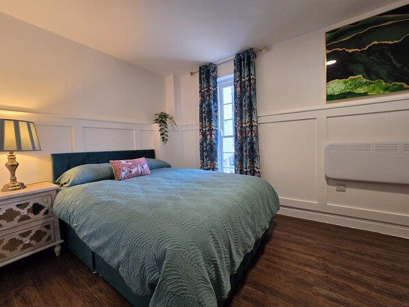 A bedroom with a bed, a nightstand, a lamp, a window with curtains, and a wall-mounted air conditioner.