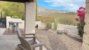 Un área de patio techada con sillas y una mesa de madera, un cactus y vistas a las montañas.