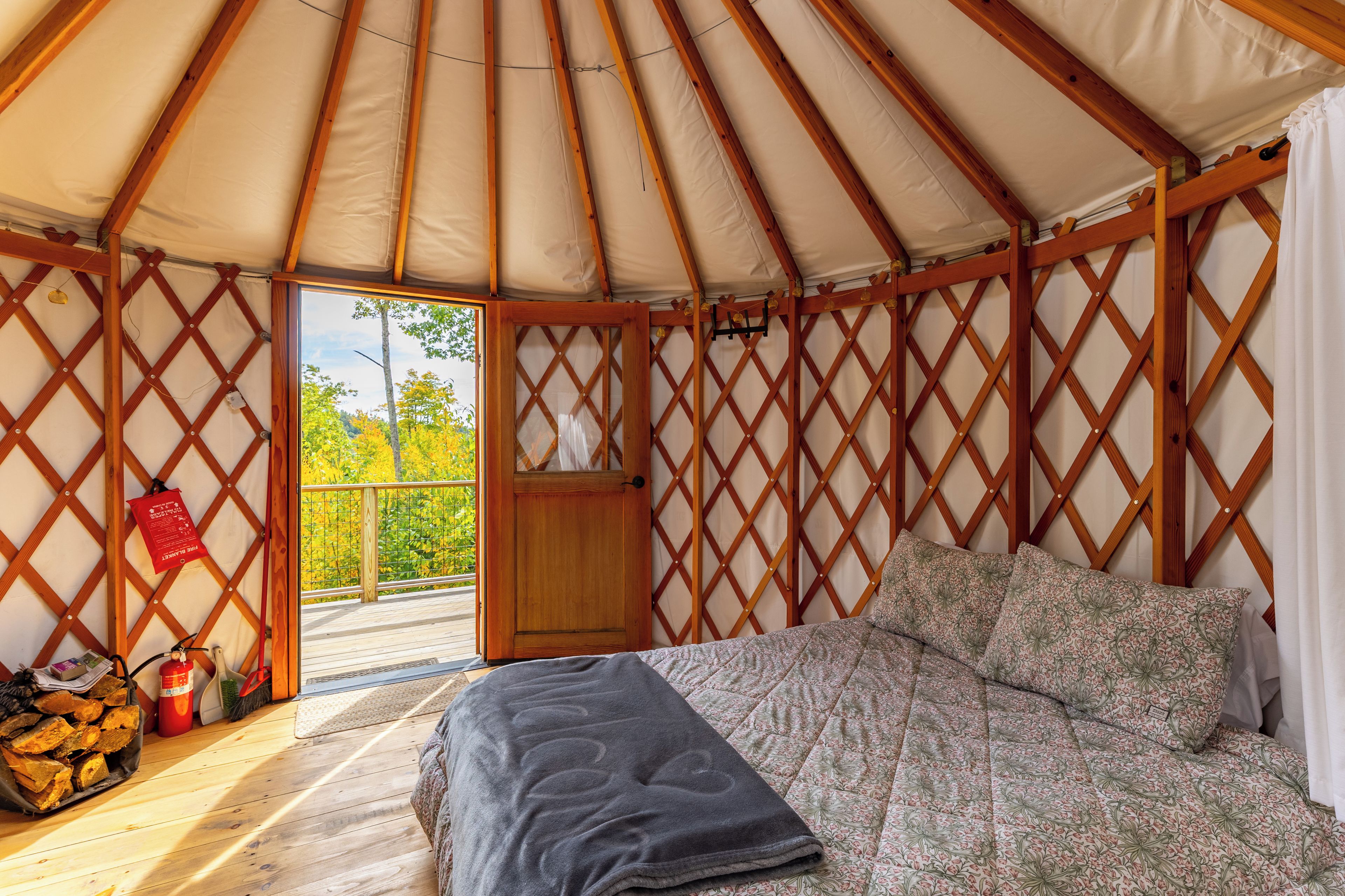 Innenansicht einer Jurte mit Bett, Wänden aus Holzgitterwerk und einer Tür, die auf einen Balkon im Freien führt.