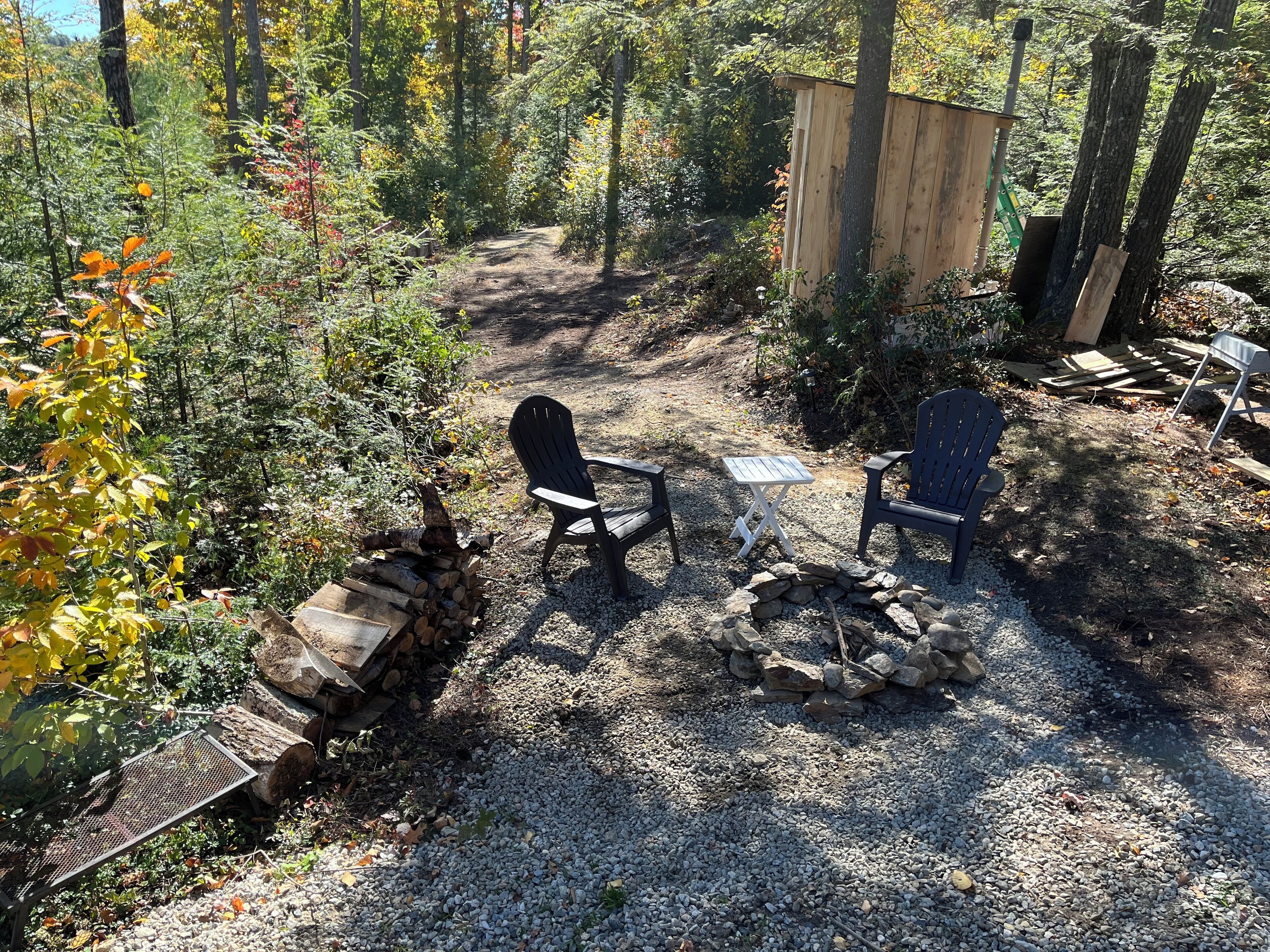 Uma área de estar rústica ao ar livre, com cadeiras Adirondack e uma pequena mesa, cercada por um caminho de cascalho e uma lareira.