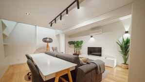 Ein modernes Wohnzimmer mit einem weißen Tisch, einer grauen Couch und einem Fernseher auf einer weißen Konsole. Auf der Konsole befinden sich Pflanzen und eine Lampe, und im Hintergrund ist eine Treppe zu sehen.