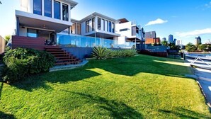 A modern two-story house with a well-maintained lawn, a glass balcony, and a clear view of the city skyline. - Luxico Bluewater Oasis (Heated Pool) (Broadbeach Waters)