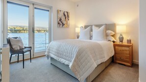 Un dormitorio con una cama grande, una silla, una mesita de noche y vistas a un lago a través de una puerta corrediza.