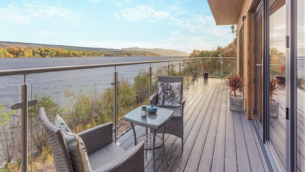 Terraza con barandilla de vidrio que da a un lago, muebles de mimbre y plantas en macetas.