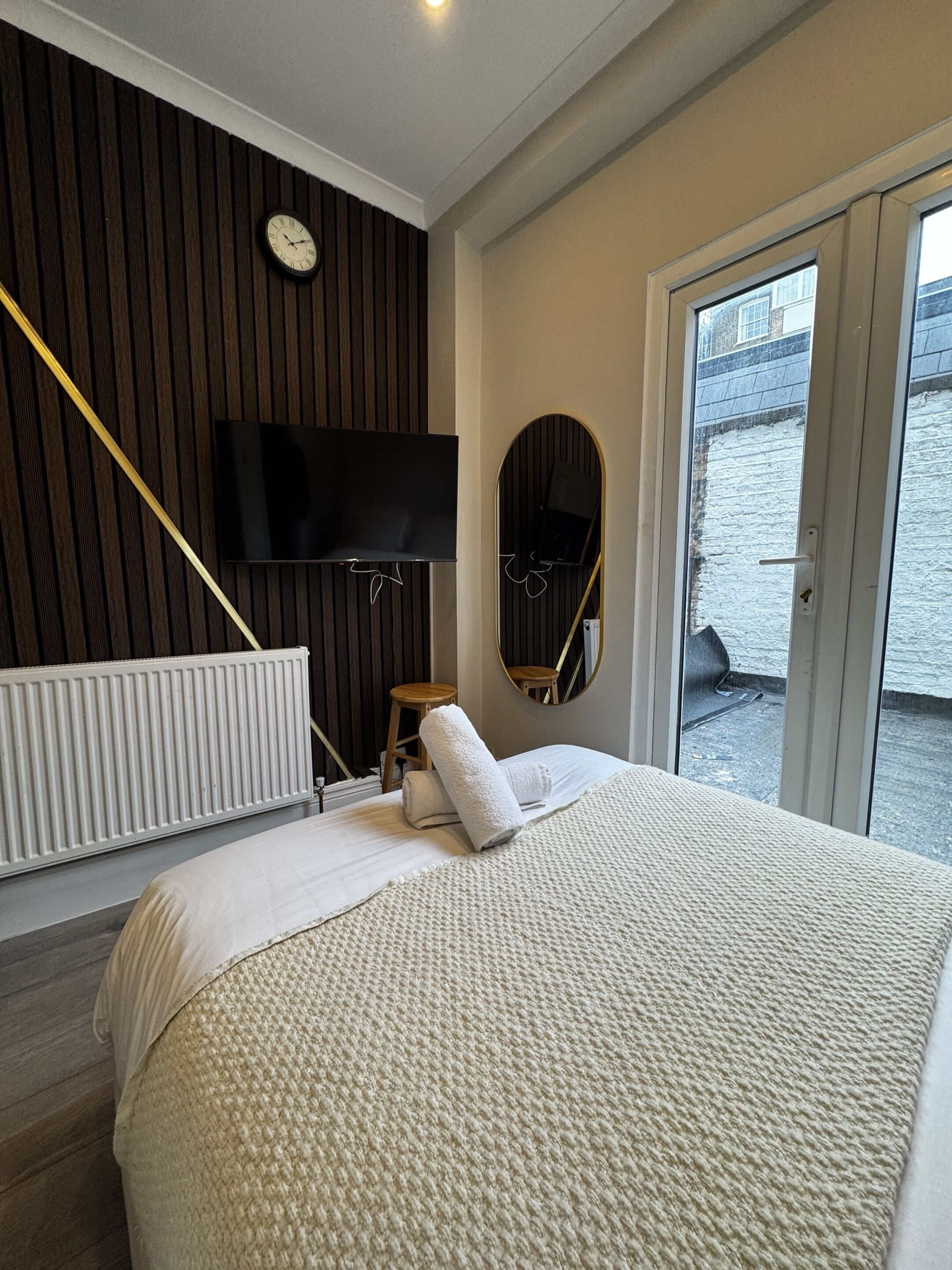 A modern bedroom with a large bed, a television mounted on a wooden panel wall, a small stool, and a mirror.
