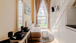 Ein kompakter Wohnbereich mit einer Couch, einem Esstisch mit schwarzen Schüsseln, einem kleinen weißen Tisch und Blick ins Freie durch große Fenster.