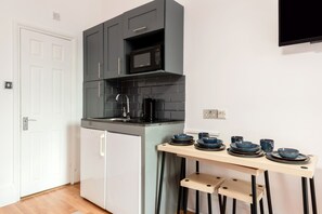 A compact kitchen with a built-in microwave, a coffee maker, and a small refrigerator. There is a wooden table with bowls and cups. - The Savers Hotel by PLI (London)