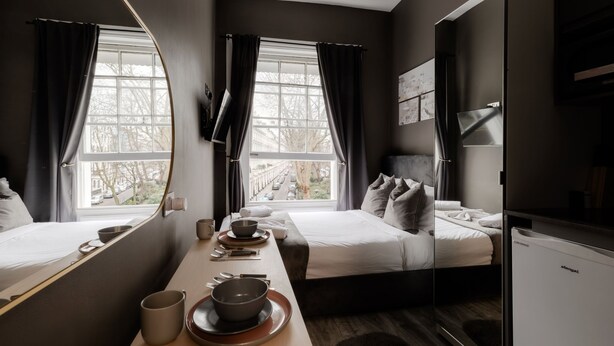 Ein modernes Hotelzimmer mit zwei Betten, einem Esstisch mit Schüsseln und Tassen sowie einem großen Fenster mit Vorhängen.