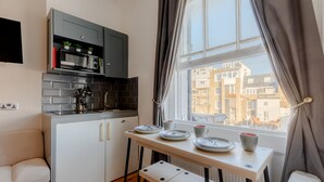 Eine kompakte Küche mit integriertem Backofen, Mikrowelle und kleinem Kühlschrank. Ein Esstisch ist mit Tellern und Tassen gedeckt, und es gibt eine Bank. Das Zimmer hat ein Fenster mit Blick auf Gebäude.