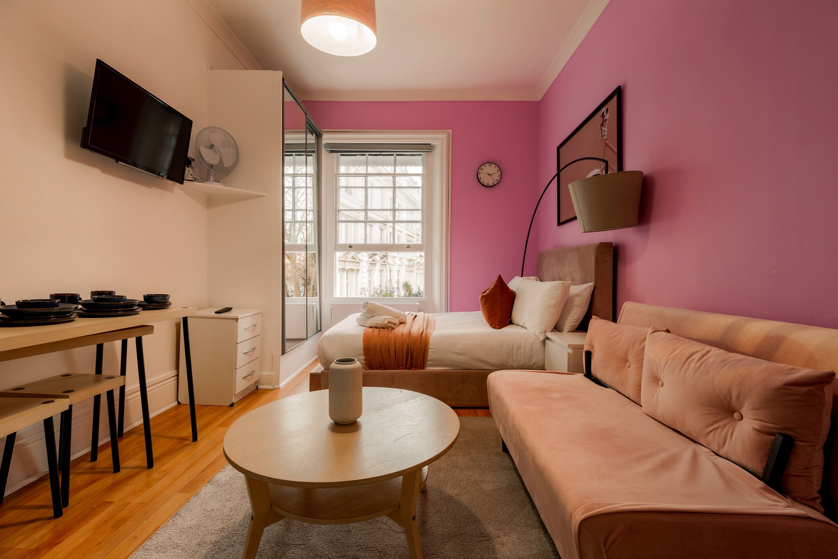 A modern living room with a pink wall, a flat-screen TV mounted above a wooden table, a sofa, and a bed with an orange throw blanket.