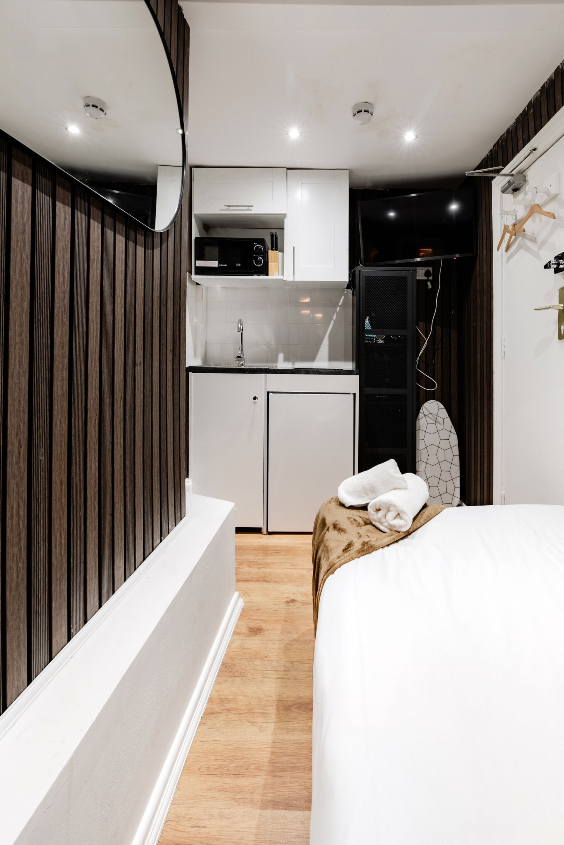 A compact hotel room with a kitchenette, a bed with towels, and a surfboard.