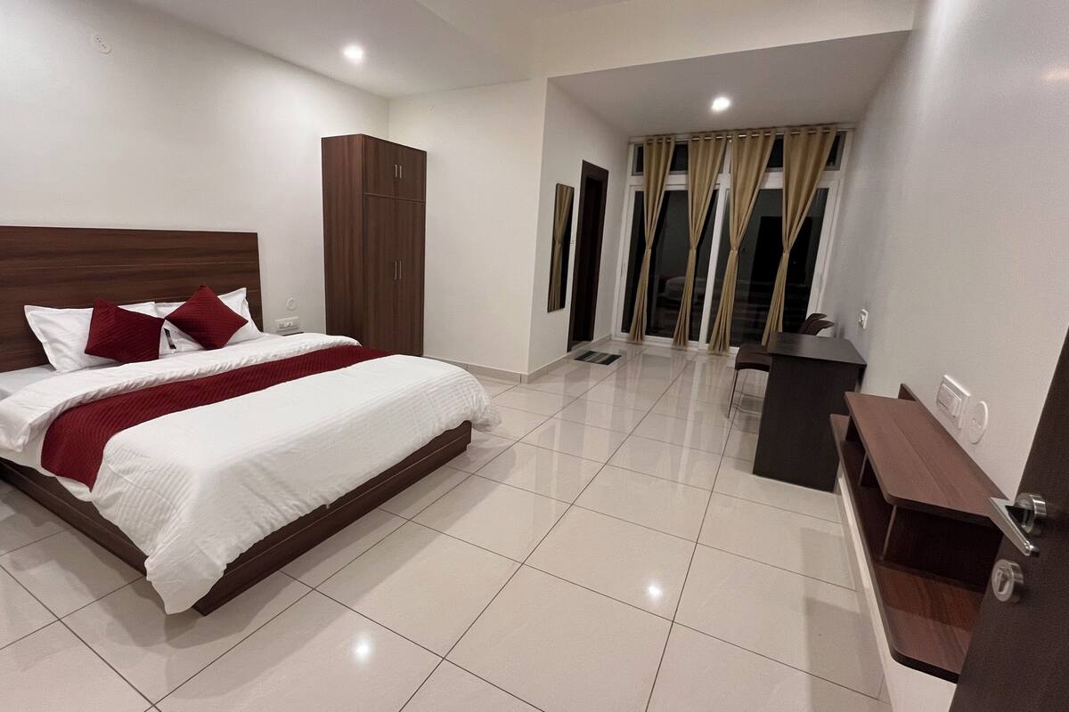 A modern hotel room with a large bed, a wardrobe, a desk, and a bathroom visible through a glass door.